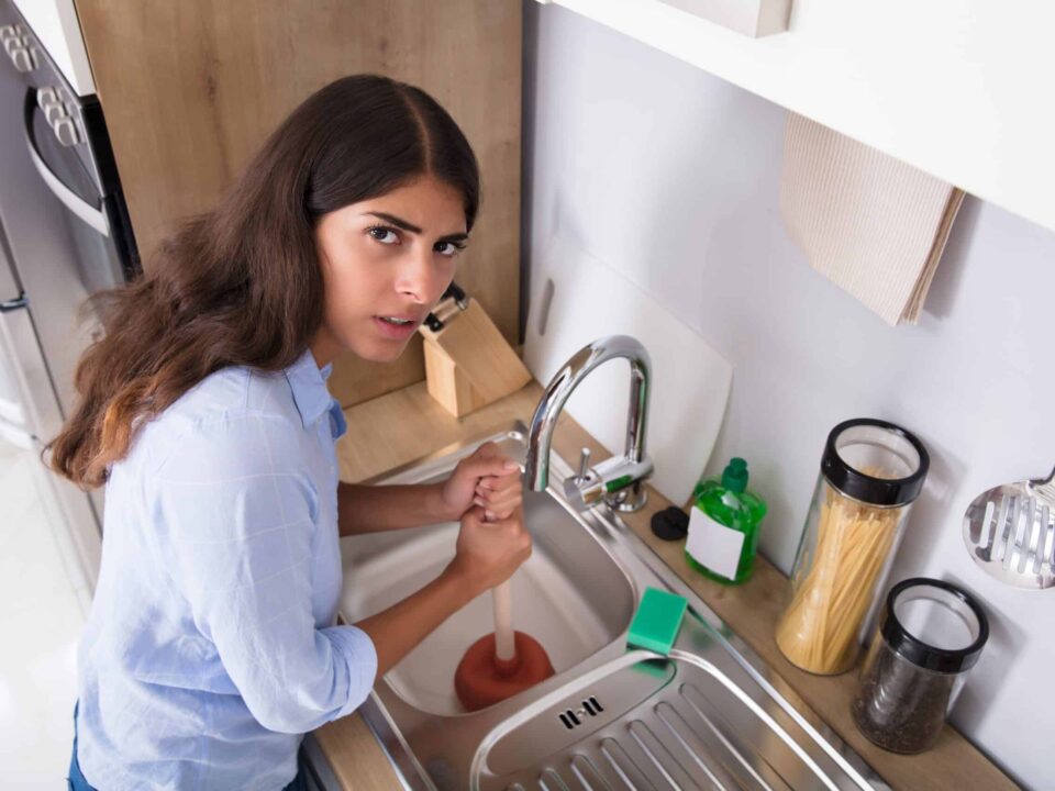 Side View Of A Young Woman Using Plunger In Blocked Kitchen Sink